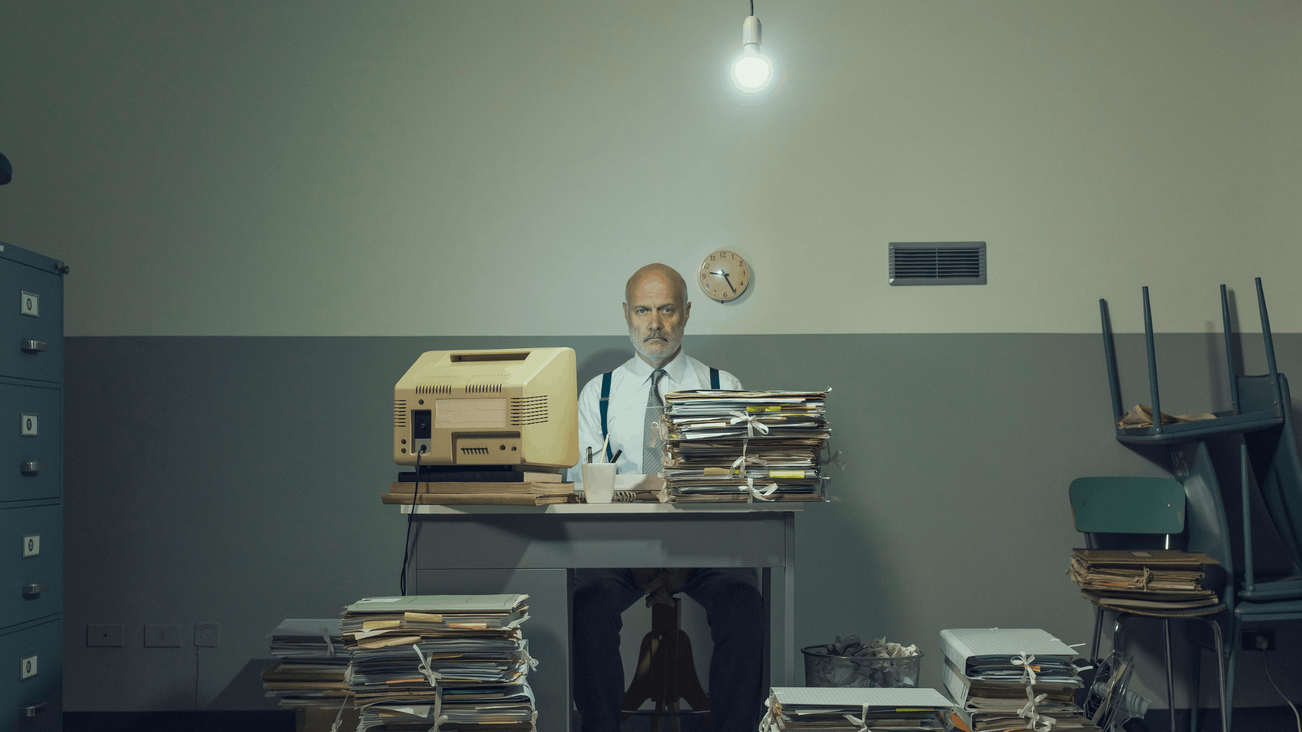 Man in suit with outdated IT solutions including old computer and files sits at desk in 70s style office
